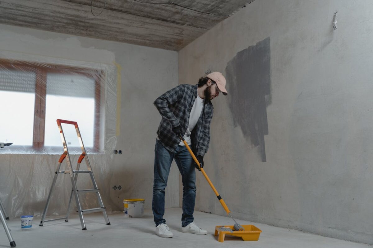 A man painting a room with a roller, using a stepladder, wearing casual attire. Renovation scene.