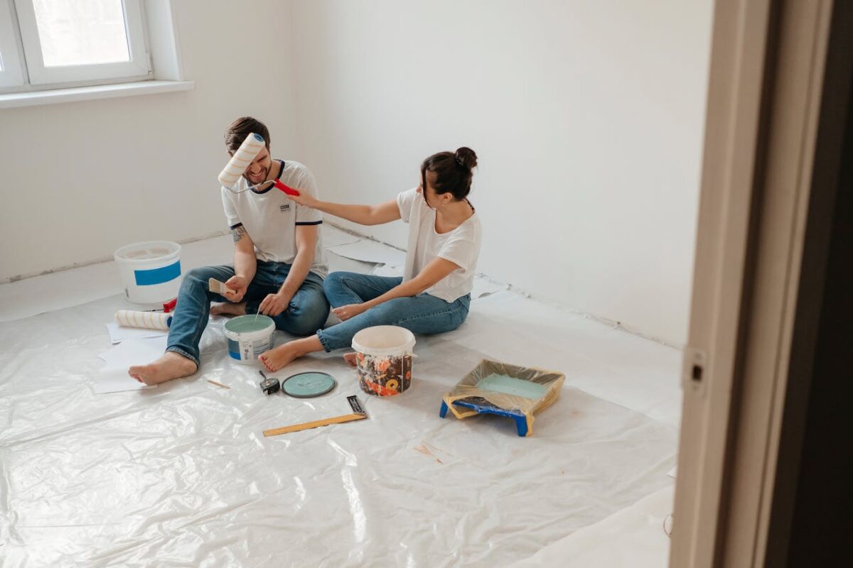 A cheerful couple sitting on the floor, painting their room with various supplies around.